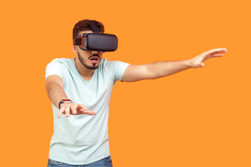 Portrait of concentrated young bearded man wearing T-shirt in virtual reality headset standing with outstretching hands. Indoor studio shot isolated on orange background. - obrazy, fototapety, plakaty