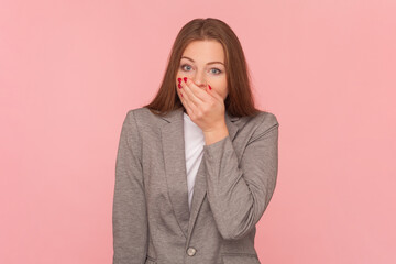 I won't say to anyone. Portrait of shocked beautiful woman with brown hair covering mouth with palm, being scared and confused, wearing business suit. Indoor studio shot isolated on pink background. - obrazy, fototapety, plakaty