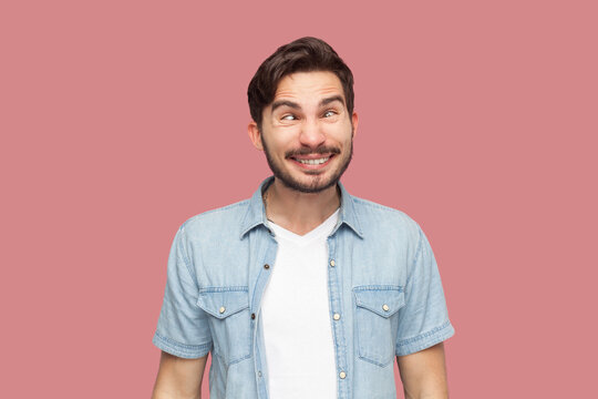 Portrait of hilarious crazy bearded man in casual style shirt standing crosses eyes, makes funny grimace, pretends to be little fool, being childish. Indoor studio shot isolated on pink background.