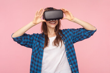 Portrait of brown haired woman in vr headset, playing virtual reality game with smile on his face, innovative technology, wearing checkered shirt. Indoor studio shot isolated on pink background.