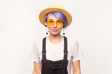 Portrait of smiling positive hipster woman with violet hair in sunglasses and hat looking at came with pleased emotion. Indoor studio shot isolated on white background.