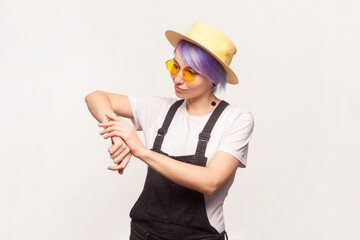 Portrait of busy serious woman with violet hair in sunglasses and hat standing looking at her smartwatch deadline. Indoor studio shot isolated on white background.