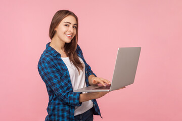 Profile portrait of smiling positive brown haired woman standing typing on laptop keyboard,remote work, distance job, wearing checkered shirt. Indoor studio shot isolated on pink background.