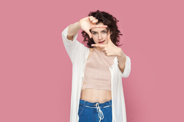 Portrait of concentrated attractive woman with curly hair wearing casual style outfit looking at camera through frame from her fingers. Indoor studio shot isolated on pink background.