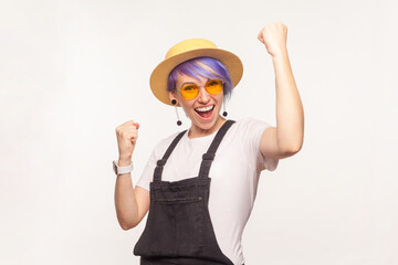 Portrait of extremely happy overjoyed woman with violet hair in sunglasses and hat clenched fists screaming with happiness celebrating her success. Indoor studio shot isolated on white background.