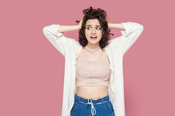 Portrait of shocked worried woman with curly hair wearing casual style outfit standing keeps hands on head, having problems, looks nervous. Indoor studio shot isolated on pink background.
