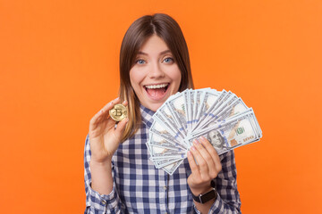 Portrait of amazed excited happy woman with brown hair showing golden bitcoin and big fan of dollar banknotes, wearing checkered shirt. Indoor studio shot isolated on orange background