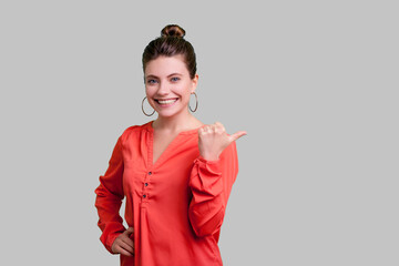 Portrait of cheerful joyful woman with bun hairstyle pointing aside at copy space for promotional text, advertisement area, wearing red blouse. Indoor studio shot isolated on gray background.