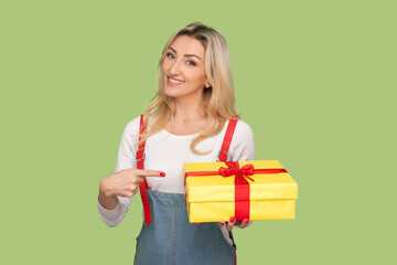 Portrait of cute happy adult blond woman holding and pointing at present box in her hand, being at birthday party, wearing denim overalls. Indoor studio shot isolated on light green background