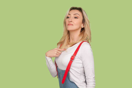 Portrait of proud selfish egoistic adult blond woman standing pointing at herself, looking at camera with arrogant face, wearing denim overalls. Indoor studio shot isolated on light green background