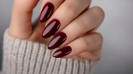 A hand holding a pair of red nails with a white sweater in the background. The nails are painted in a deep burgundy color