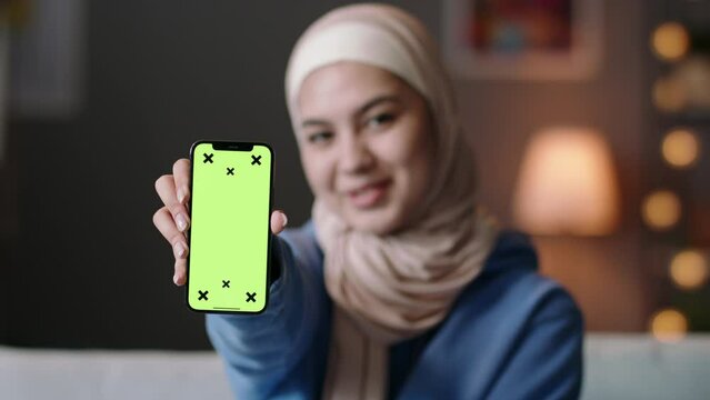 Woman shows phone with green screen. Young muslim woman holds smartphone, shows green chroma key to camera on phone display. Template for embedding videos, photos, ads, web pages, online phone apps