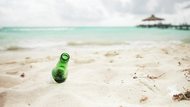 Close up of an empty green glass beer bottle stuck in the white sand on the beach. Turquoise sea waves and pier with gazebo in the background. People litter in wildlife nature and pollute ecosystem