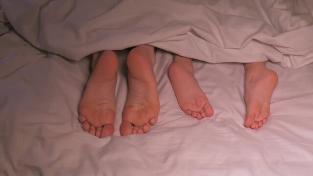 dancing legs of boy son child and mother female foot lie under the blanket on sheet in the evening. Happy baby kid girl daughter and woman mom wiggling bare feet in bed in the bedroom