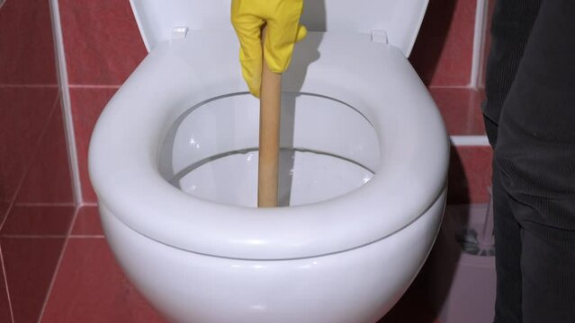Cleaning blockages in the toilet indoor. A woman in gloves doing cleaning work with plunger in the toilet bowl.