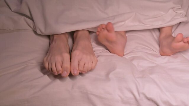 dancing legs of boy son child and mother female foot under blanket on sheet in the evening. Happy baby kid girl daughter and woman mom wiggling bare feet in bed in the bedroom