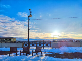 Serene Winter Sunset at Snowy Riverfront With Benches and Streetlamp. Sun sets over a tranquil,...