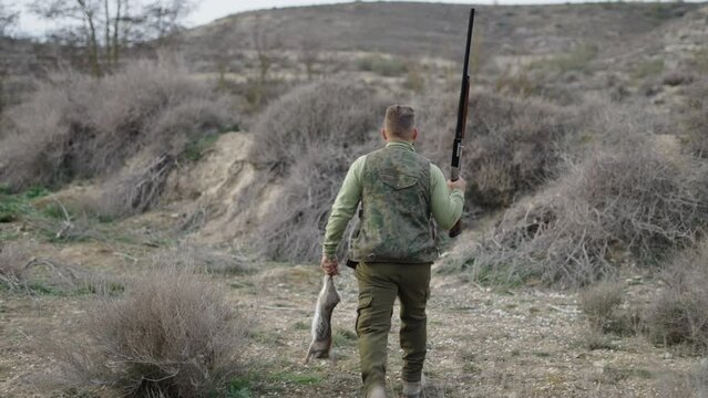 A hunter walks backwards with a rifle and a rabbit