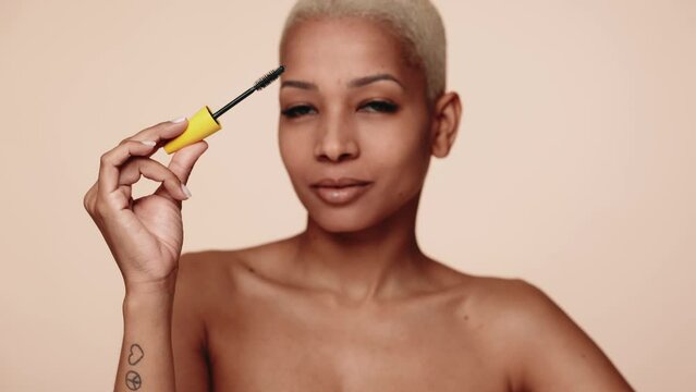 A poised Hispanic woman with short hair confidently showing mascara brush, showcasing a daily makeup routine against a neutral backdrop.