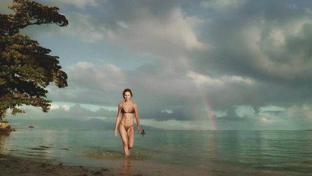 A woman wearing a bikini is walking along the sandy shore of the beach.