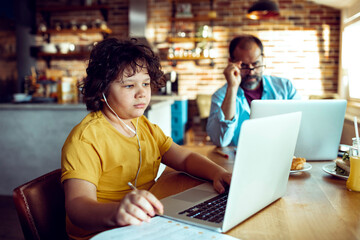 Happy child doing homework on laptop while father works in background at home