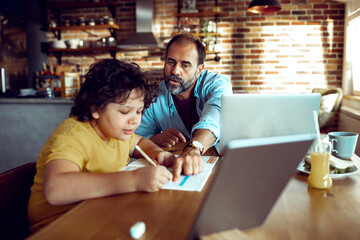 Happy child doing homework on laptop while father works in background at home