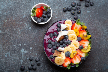 Healthy summer acai smoothie bowl with chia seeds, fresh banana, strawberry, blueberry, cocos, kiwi top view on rustic concrete background with spoon - 769948627