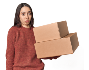 Caucasian young woman with moving boxes on studio background shrugs shoulders and open eyes confused.