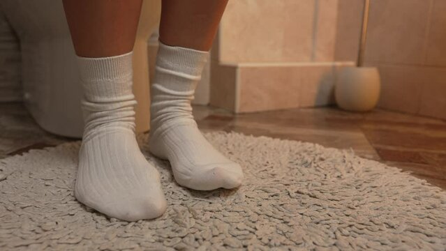 Camera smoothly zooms in on women's feet in white warm socks on a white fleecy toilet mat, girl tapping her toes on the floor