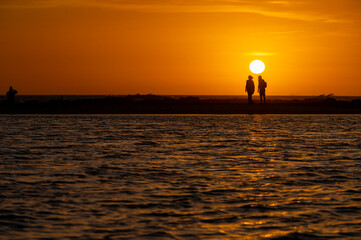 Man and woman staying and looking at sunset on ocean beach, orange sky, silhouettes of people on...