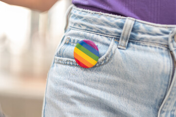 A person is wearing a blue pair of jeans and has a rainbow pin on their pocket