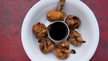 Top view of deep fried breaded chicken wings with a bittersweet dipping sauce, in a white bowl on the red wooden table.