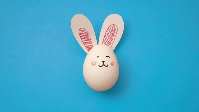 White chicken egg with cute bunny face and rabbit ears on blue background. Top view 4K animated video. Magic frame-by-frame stop motion animation for Happy Easter. Postcard for religious holiday. Gift
