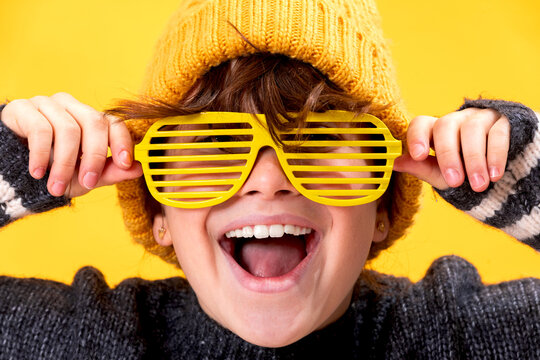 A gleeful young girl lifts her funky yellow shutter shades while laughing, with a matching yellow beanie against a monochrome background