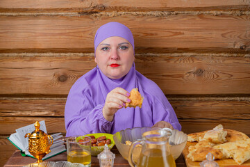 A Muslim woman in a lilac hijab at the table eats lamb with flatbread on the holiday of breaking...