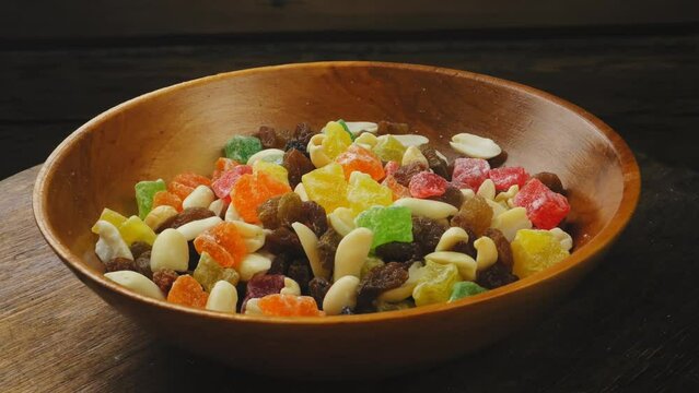 Mix of multi-colored dried fruits, candied fruits and scattered nuts in a wooden plate on a black background, rotating in a circle. Medium plan