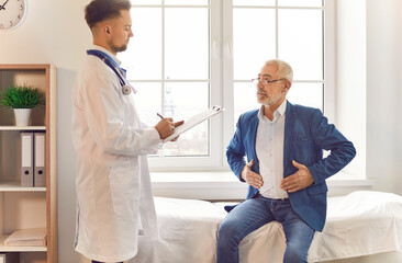 Doctor consulting male senior patient talking, sharing stomach symptoms, belly complaining, anxious, troubled about health. Old man visiting clinic for medical care, treatment plan, asking for help - obrazy, fototapety, plakaty