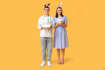Happy young couple with Easter bunny ears, basket and cake on yellow background