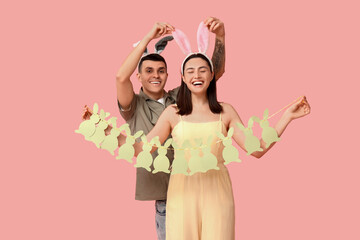 Happy young couple with Easter garland and bunny ears on pink background - obrazy, fototapety, plakaty