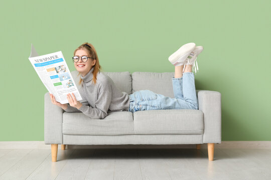 Happy young woman with newspaper lying on sofa near green wall