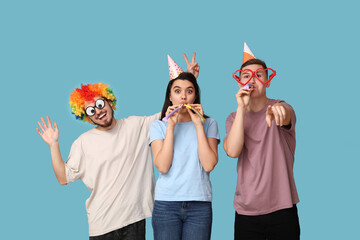 Young friends in funny disguise with party whistles pointing at viewer on blue background. April...