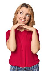 Blonde middle-aged Caucasian woman in studio keeps hands under chin, is looking happily aside.