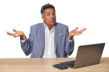Elegant businessman at desk with laptop doubting and shrugging shoulders in questioning gesture.