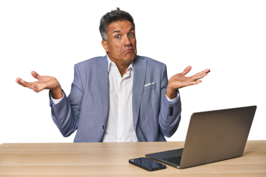 Elegant businessman at desk with laptop doubting and shrugging shoulders in questioning gesture.