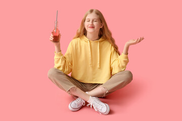 Beautiful young woman with bottle of soda sitting against pink background