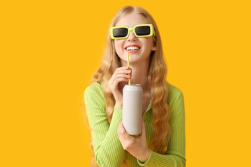 Happy young woman in sunglasses with can of soda on yellow background