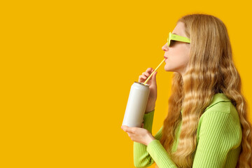Beautiful young woman in sunglasses with can of soda on yellow background