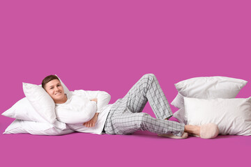 Young man in pajamas with pillows lying on purple background