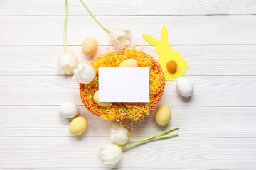 Basket with blank card, Easter eggs and tulips on white wooden background