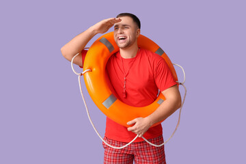 Happy young lifeguard with whistle and lifebuoy looking at somewhere on lilac background